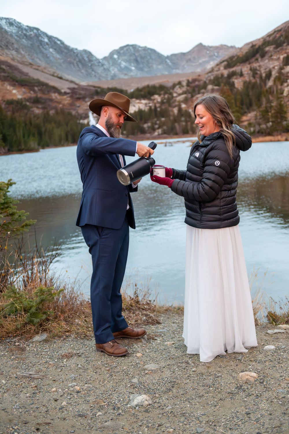 A groom pours coffee to his bride at sunrise before their elopement at a high mountain lake with rugged peaks surrounding them.