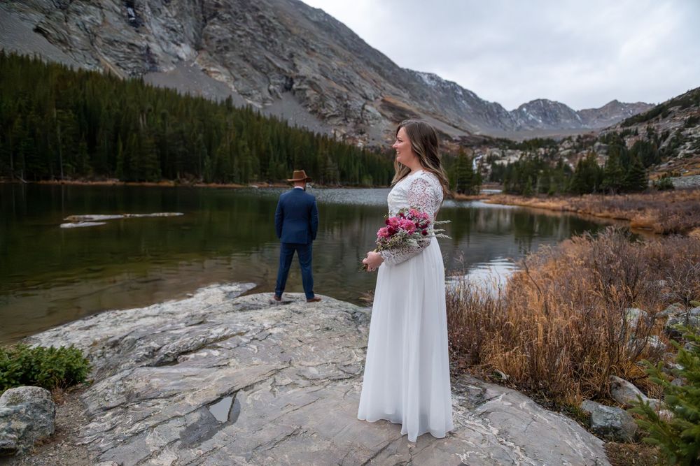 Bride holding pink bouquet stands on rocky shore with groom overlooking mountain lake and pine forest backdrop.