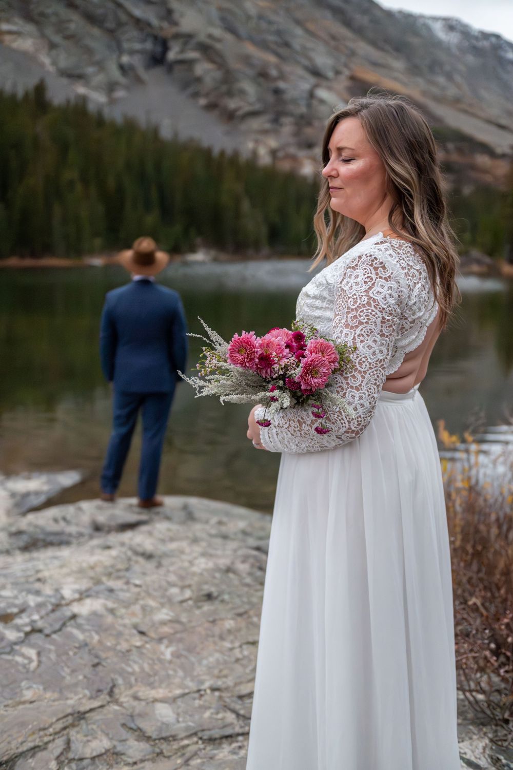 A bride in a white lace dress stands behind her groom  in a blue suit on a rocky outcropping. She is holding a pink wildflower bouquet.