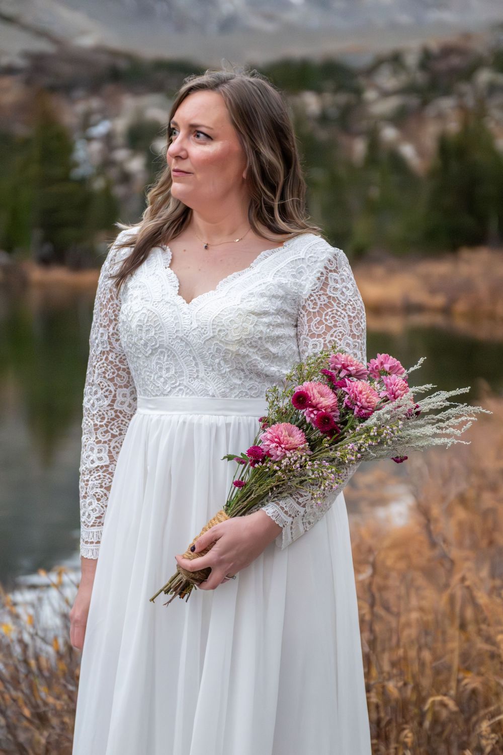 A bride in white lace long-sleeve wedding gown holding pink bouquet outdoors near mountain lake in autumn.