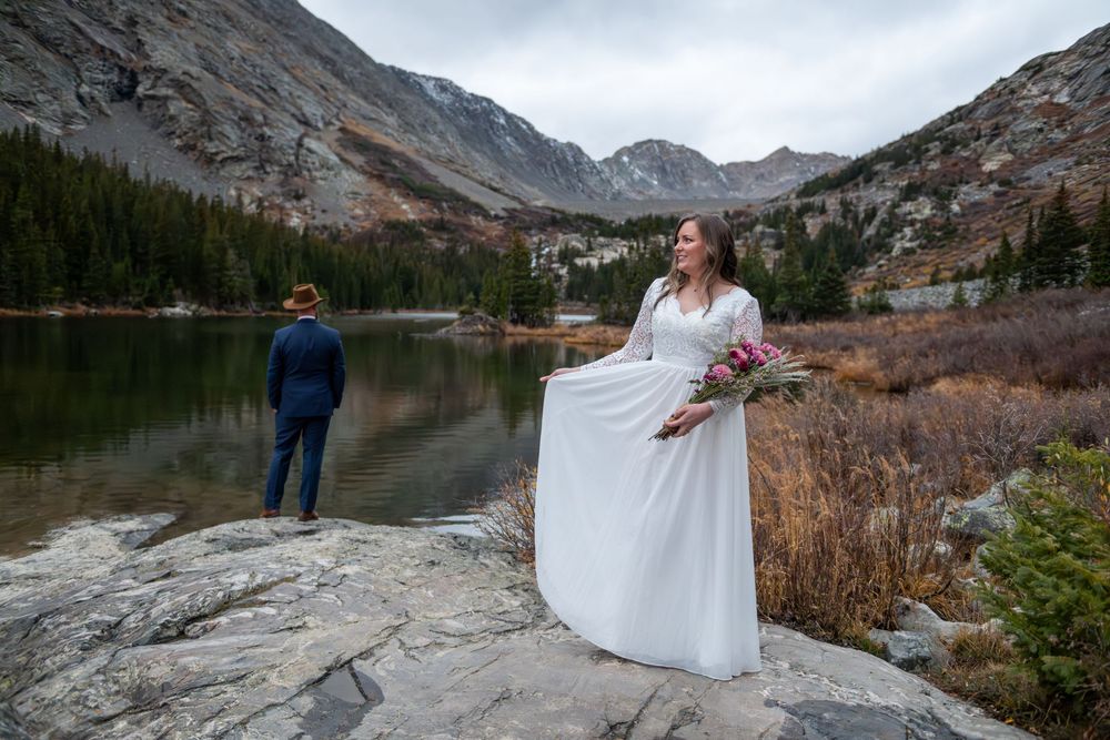 A bride in white gown holds bouquet near a mountain lake. The groom in blue suit stands at the water's edge with a brown cowboy hat on.