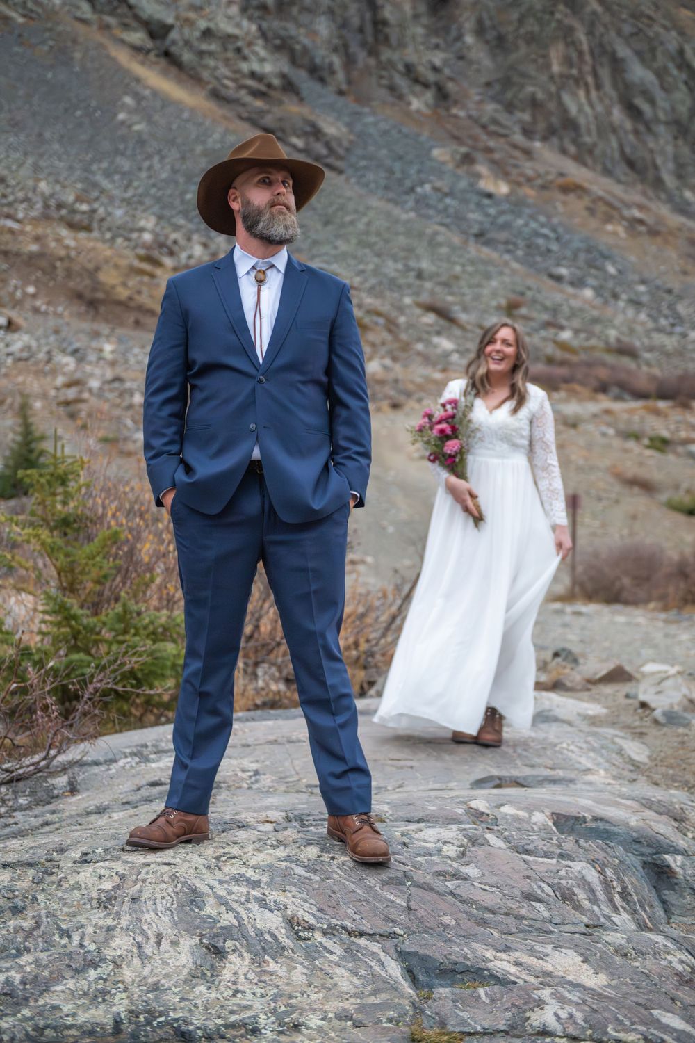 A bride stands behind her groom before her first look on a rocky outcropping in the mountains. She wears a white lace dress, he a blue suit.