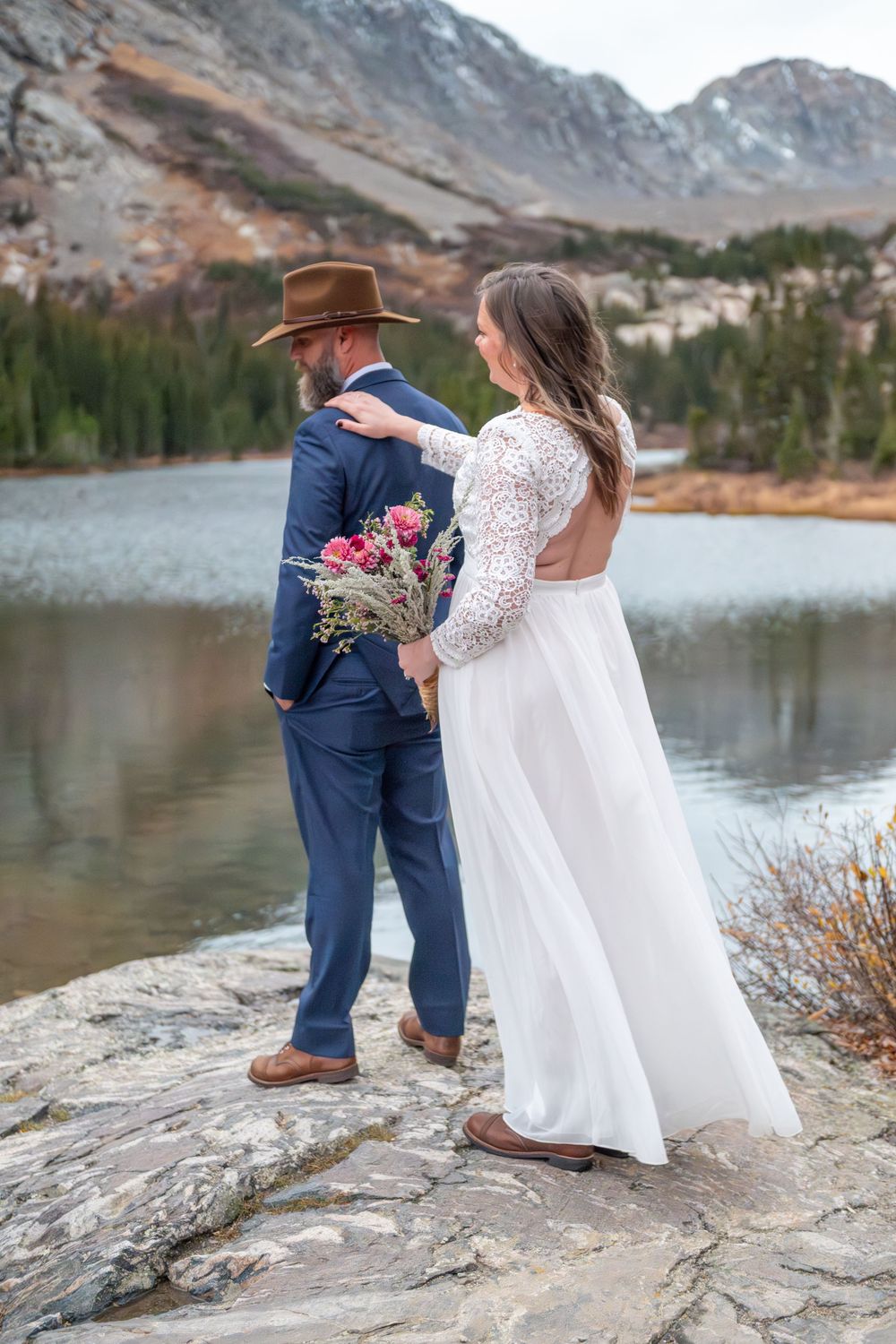 Bride and groom standing on rocky shore of mountain lake, woman in white lace dress holding pink bouquet, man in blue suit and brown hat.