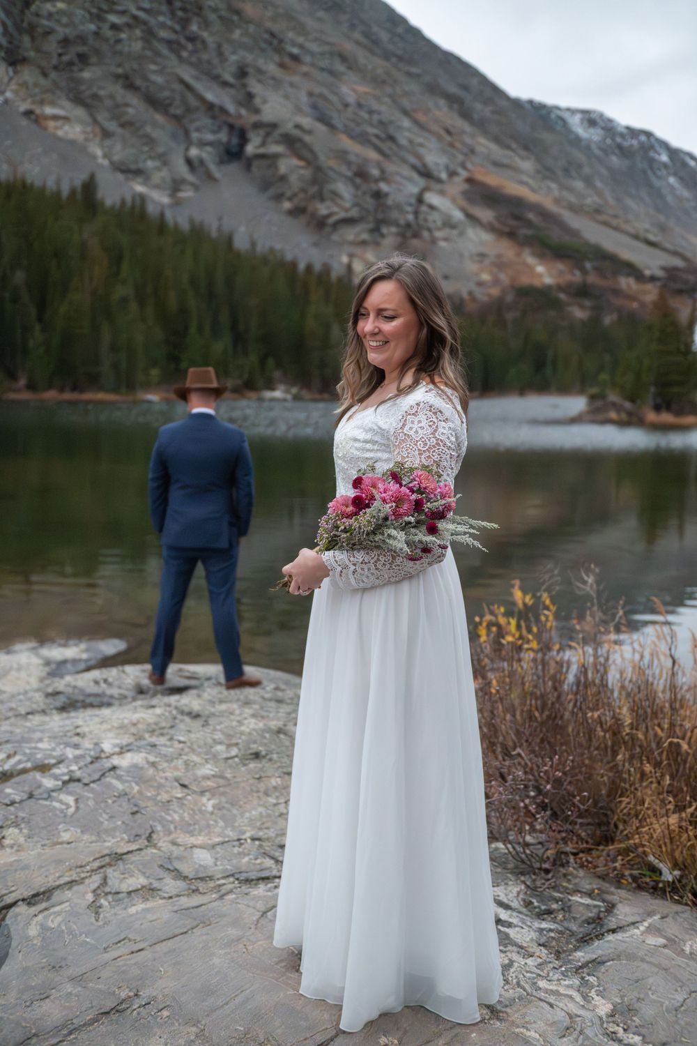 Bride in white lace gown holds a pink bouquet by mountain lake, groom in navy suit stands behind her at the edge of the lake.