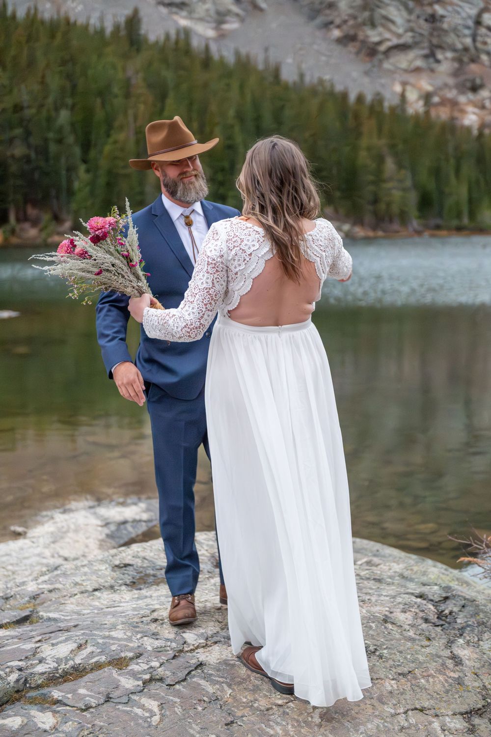 A groom smiles at his bride. they are facing each other. She is holding a pink wildflower bouquet. He wears a brown, felt cowboy hat.