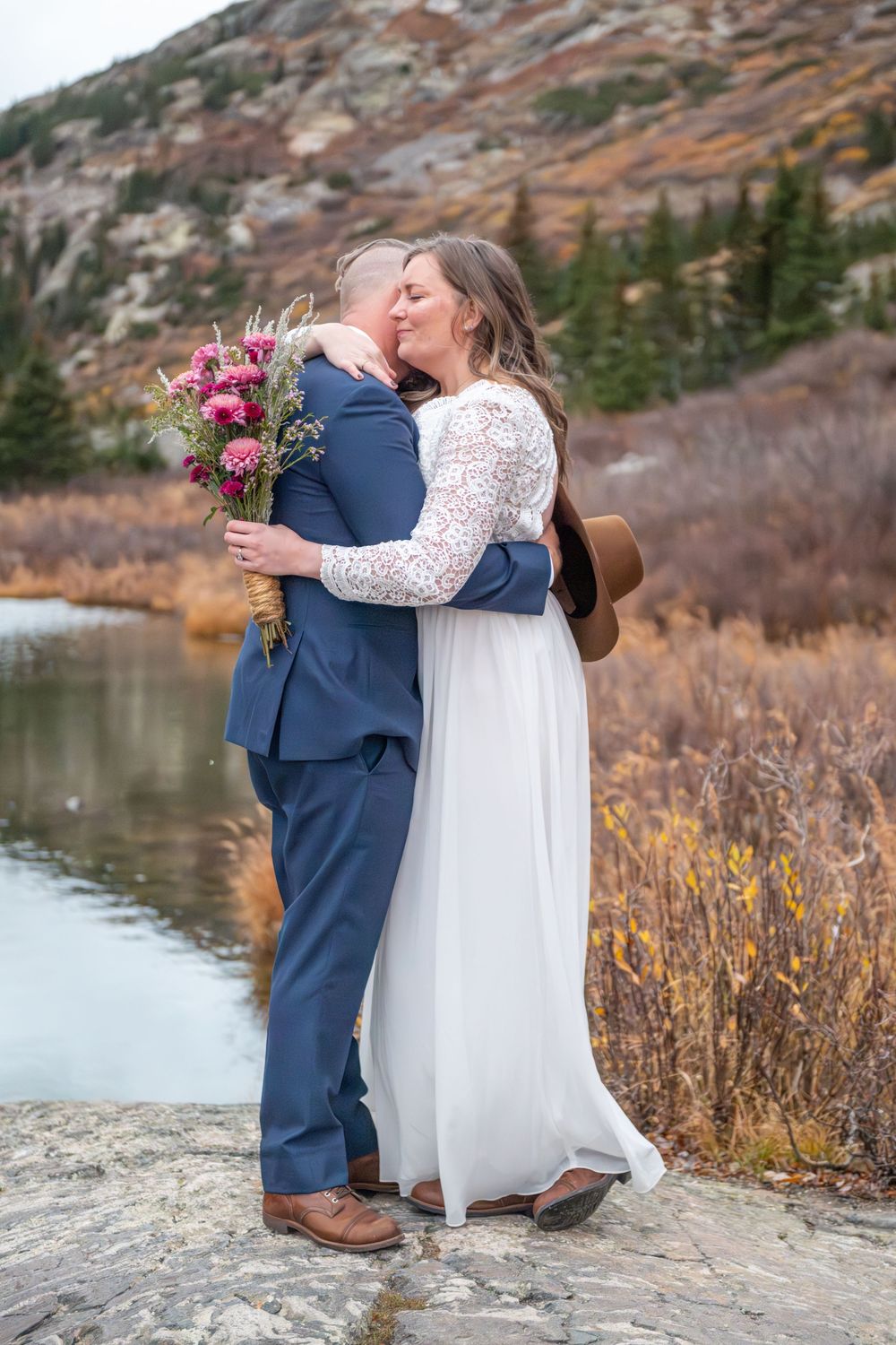 An eloping couple shares a romantic embrace outdoors during their fall wedding elopement, bride holding a pink bouquet by a mountain lake.