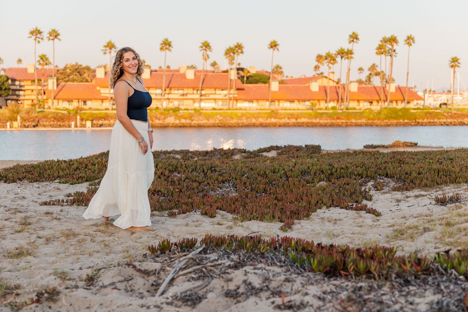 A romantic couple in white attire walks along a scenic beach at sunset with palm trees in the background.