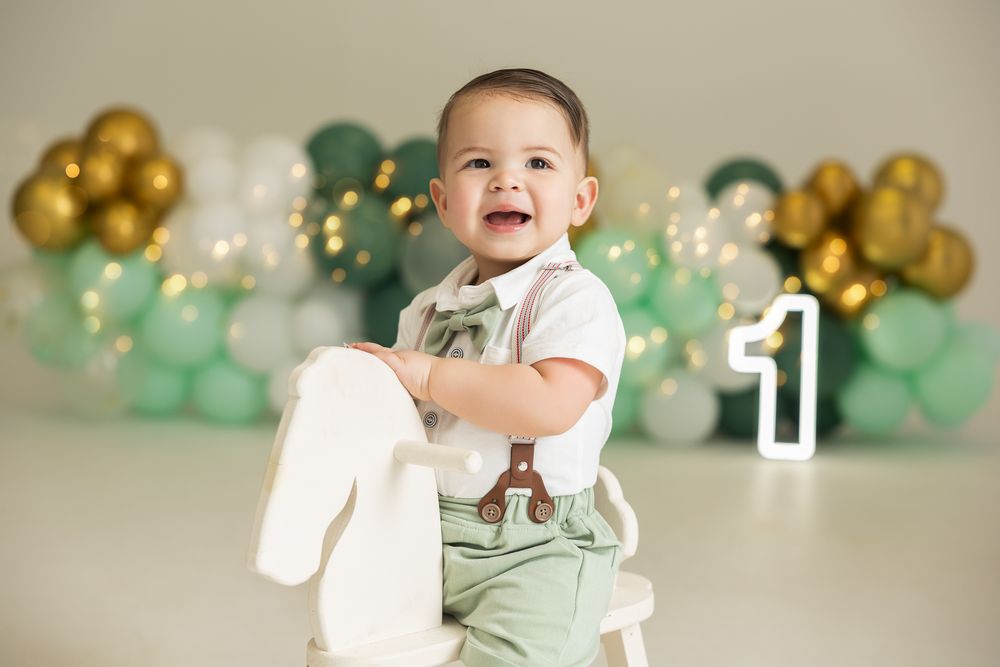 Smiling baby boy in suspenders sits on rocking horse with green and gold balloon backdrop and number one sign.
