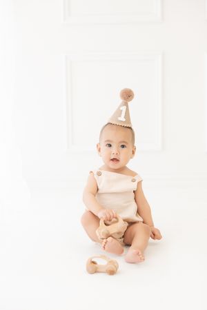 Cute baby in beige romper wearing a number 1 birthday hat sits on white floor with a wooden toy car.