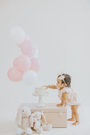 Toddler girl in pink dress reaching for white cake on stand, surrounded by bunny toys and pink balloons.
