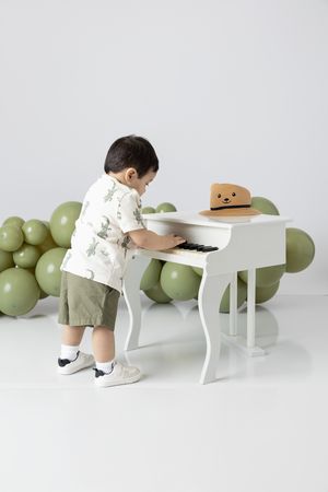 Small child playing white piano with green balloons and teddy bear decoration.