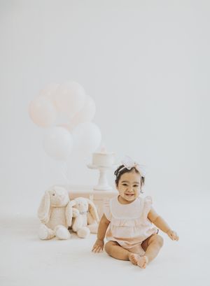 Baby girl in pink romper sits smiling beside bunny toys and white balloons on a white backdrop.