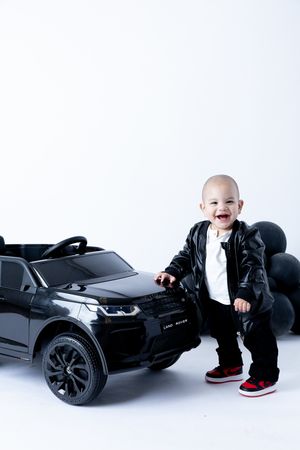 Toddler in leather jacket poses beside black toy luxury car.