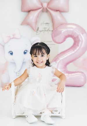 Toddler girl in white dress sits smiling at her 2nd birthday party with pink balloons and elephant decor.