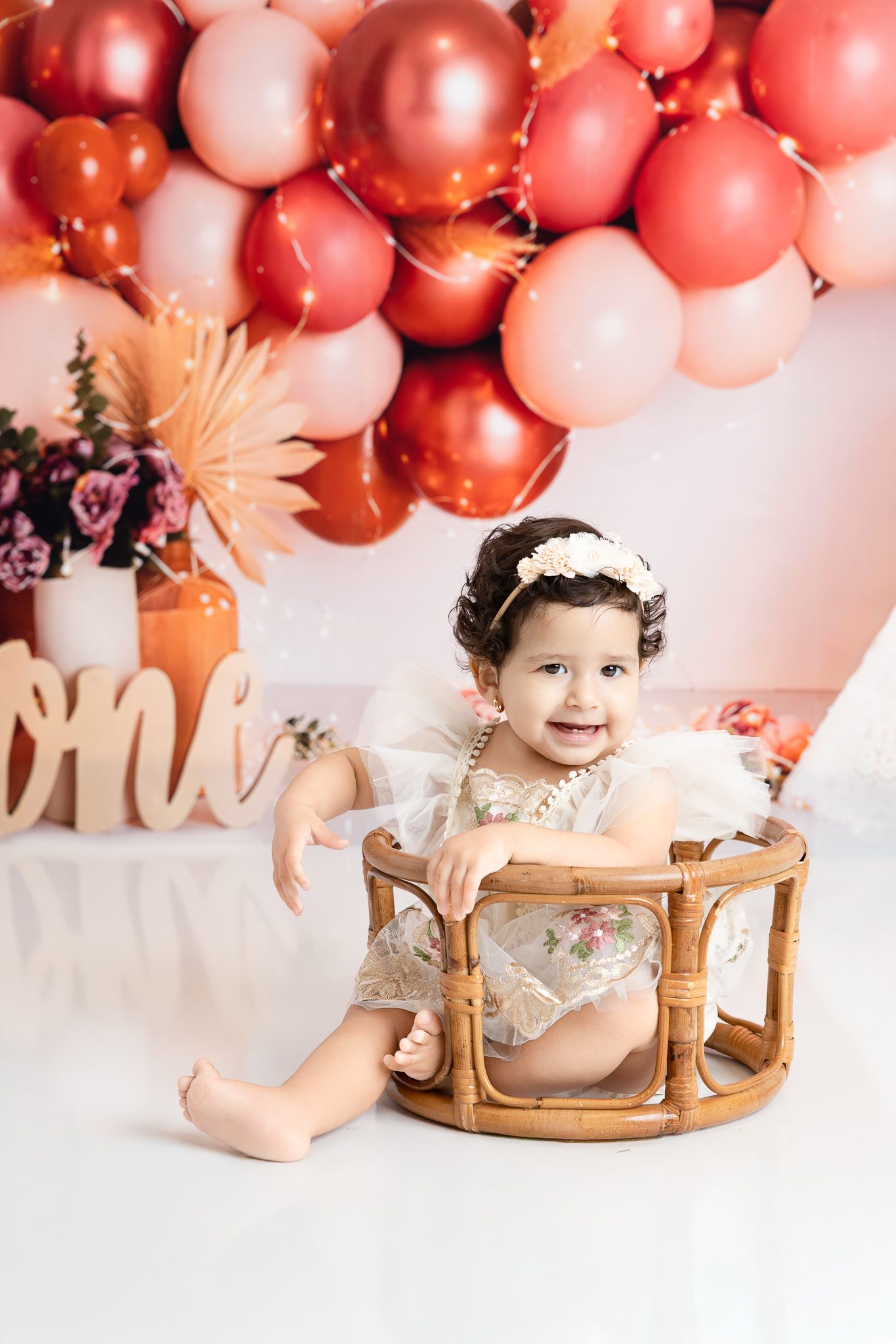 Baby sitting in wicker chair with pink and rose gold balloon arch decoration.
