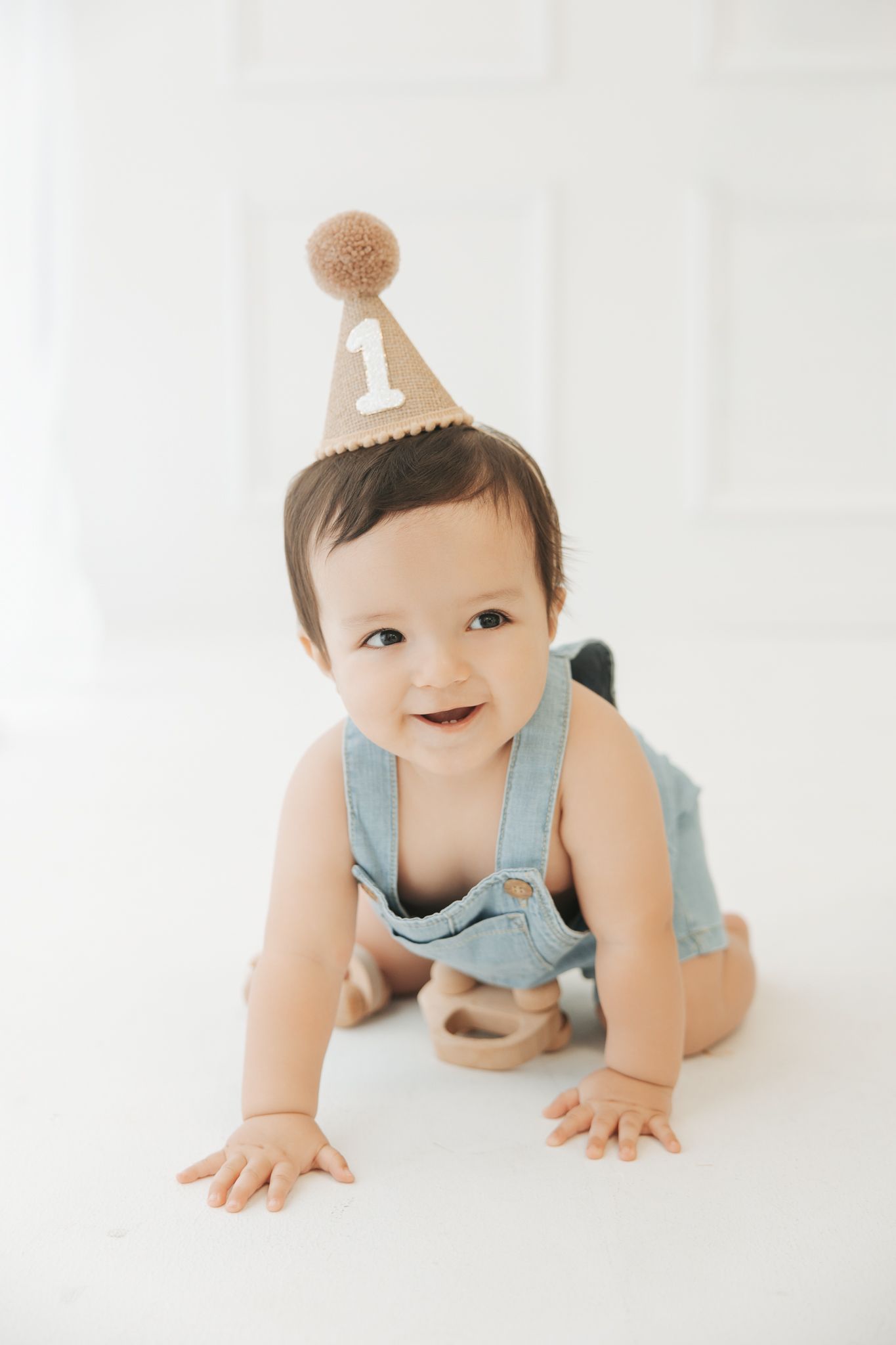 Smiling baby in denim overalls wearing a number one birthday party hat crawling on white floor.