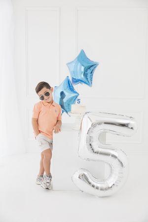 Boy in sunglasses celebrating 5th birthday with silver number balloon and blue star balloons in white room.