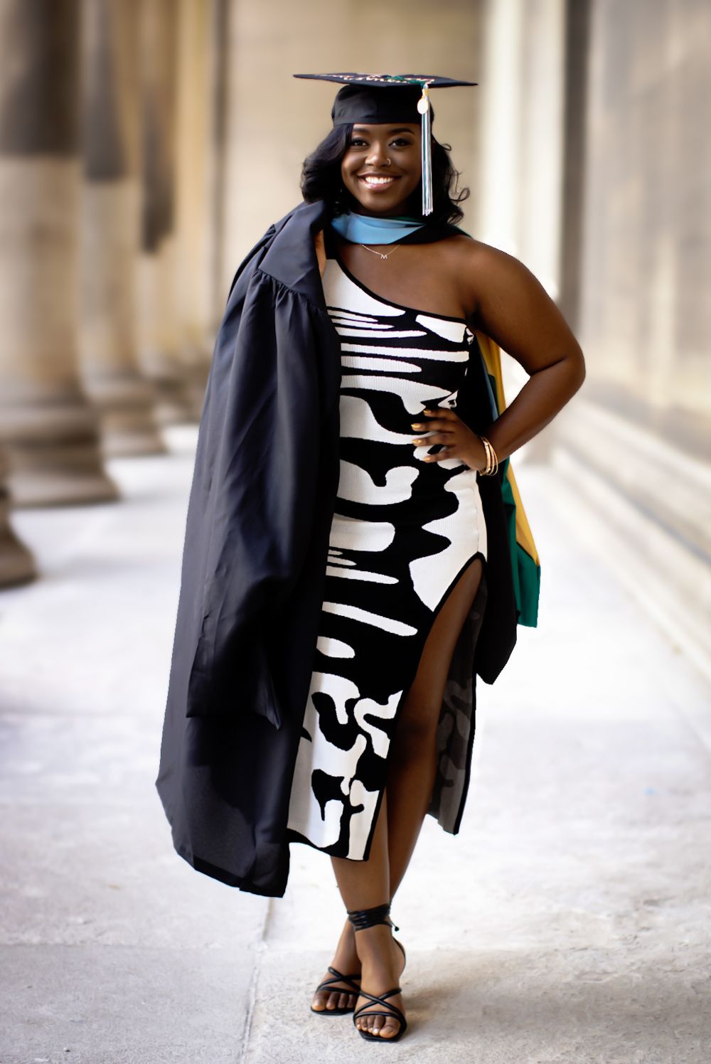 A graduate in a black cap and gown poses with a black and white striped dress for graduation photos in a hallway.