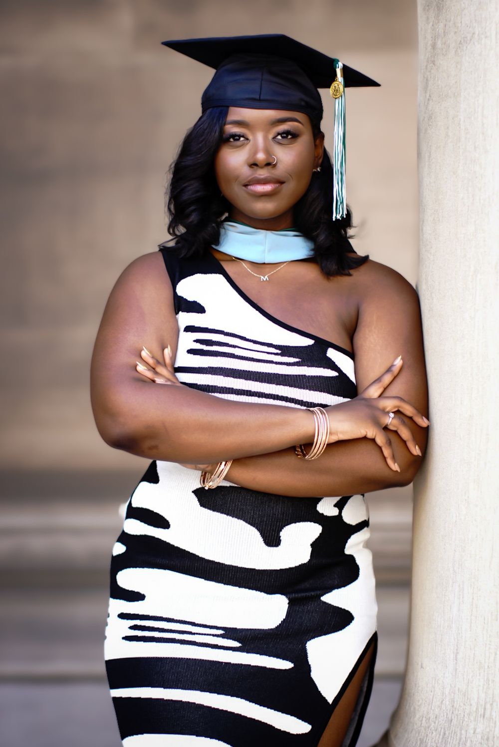 Graduate in black and white dress poses confidently against light colored pillar wearing graduation cap and tassel.