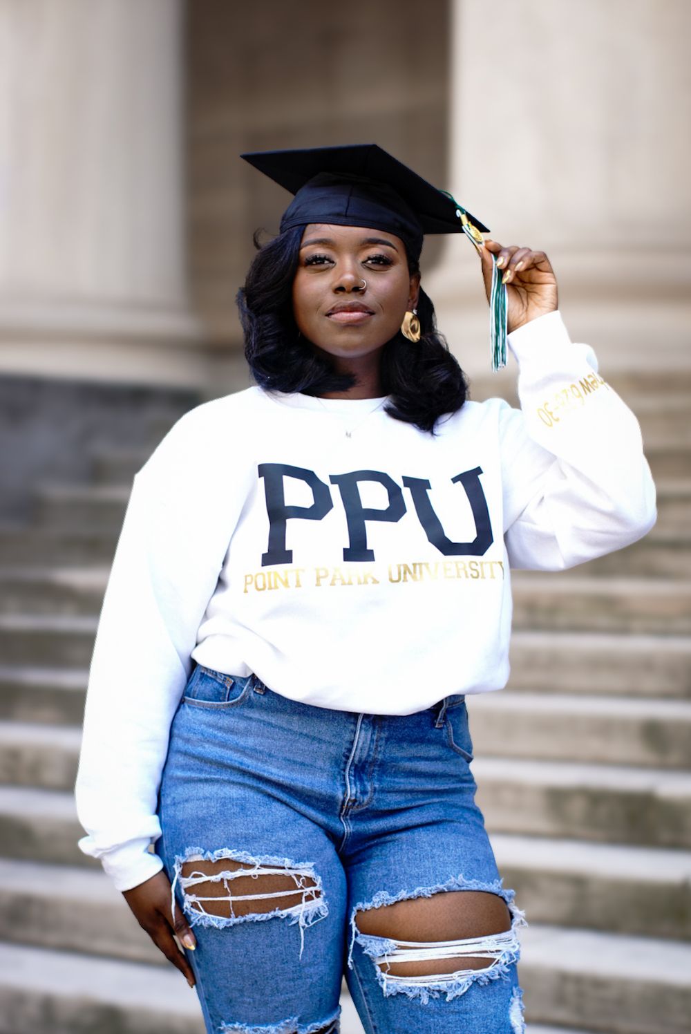 Graduate in cap and gown wearing PPU sweatshirt and ripped jeans poses on stone steps for graduation photo.