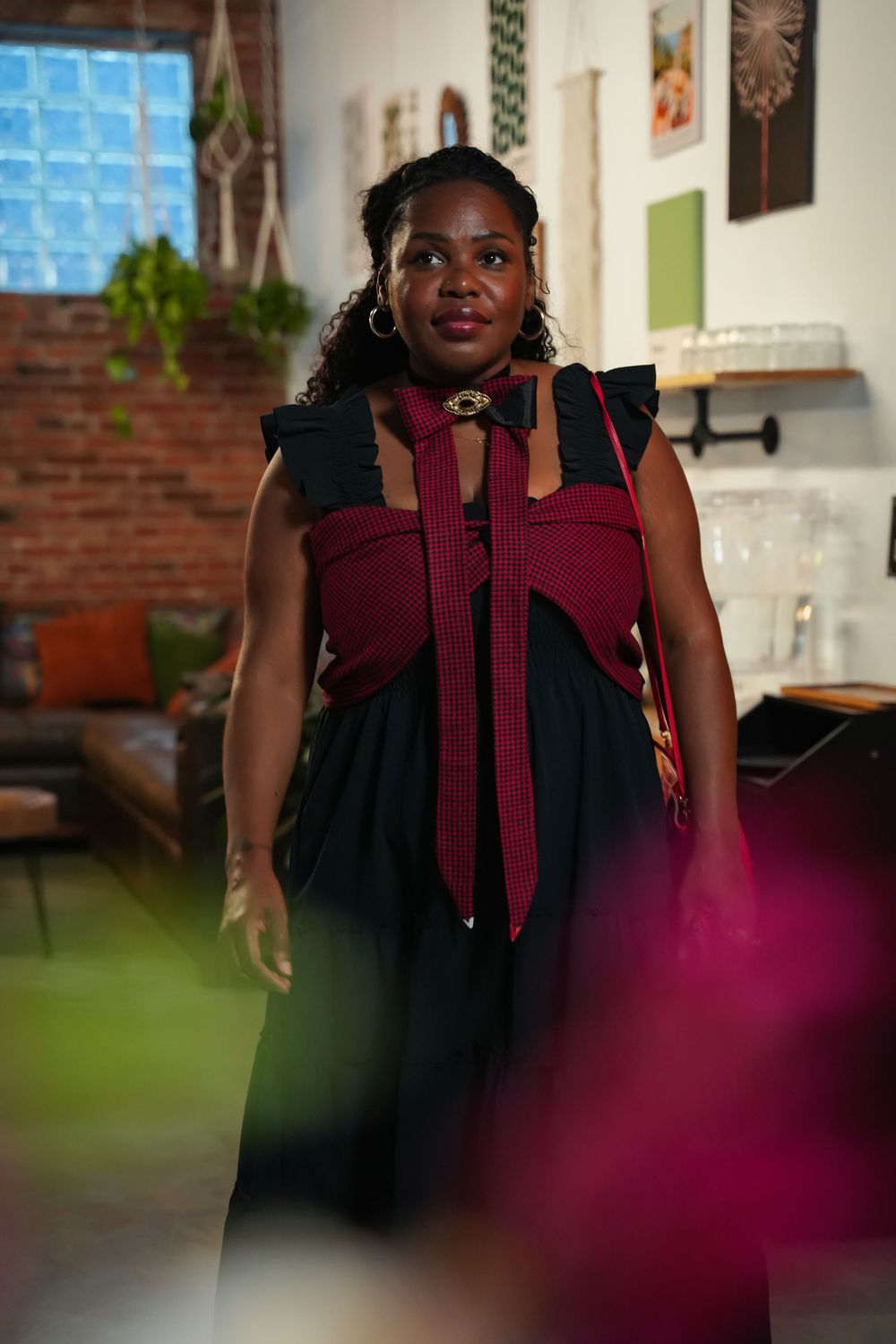 A person in a burgundy dress with a bow detail stands in an indoor setting with brick walls and artwork.
