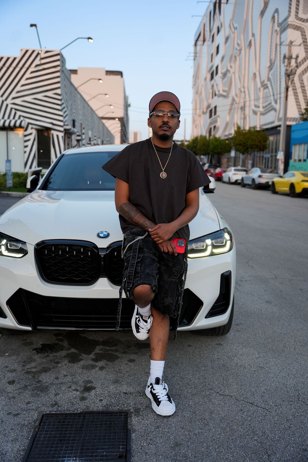 Person in casual streetwear poses in front of white BMW M4 on urban street with muraled buildings in background.