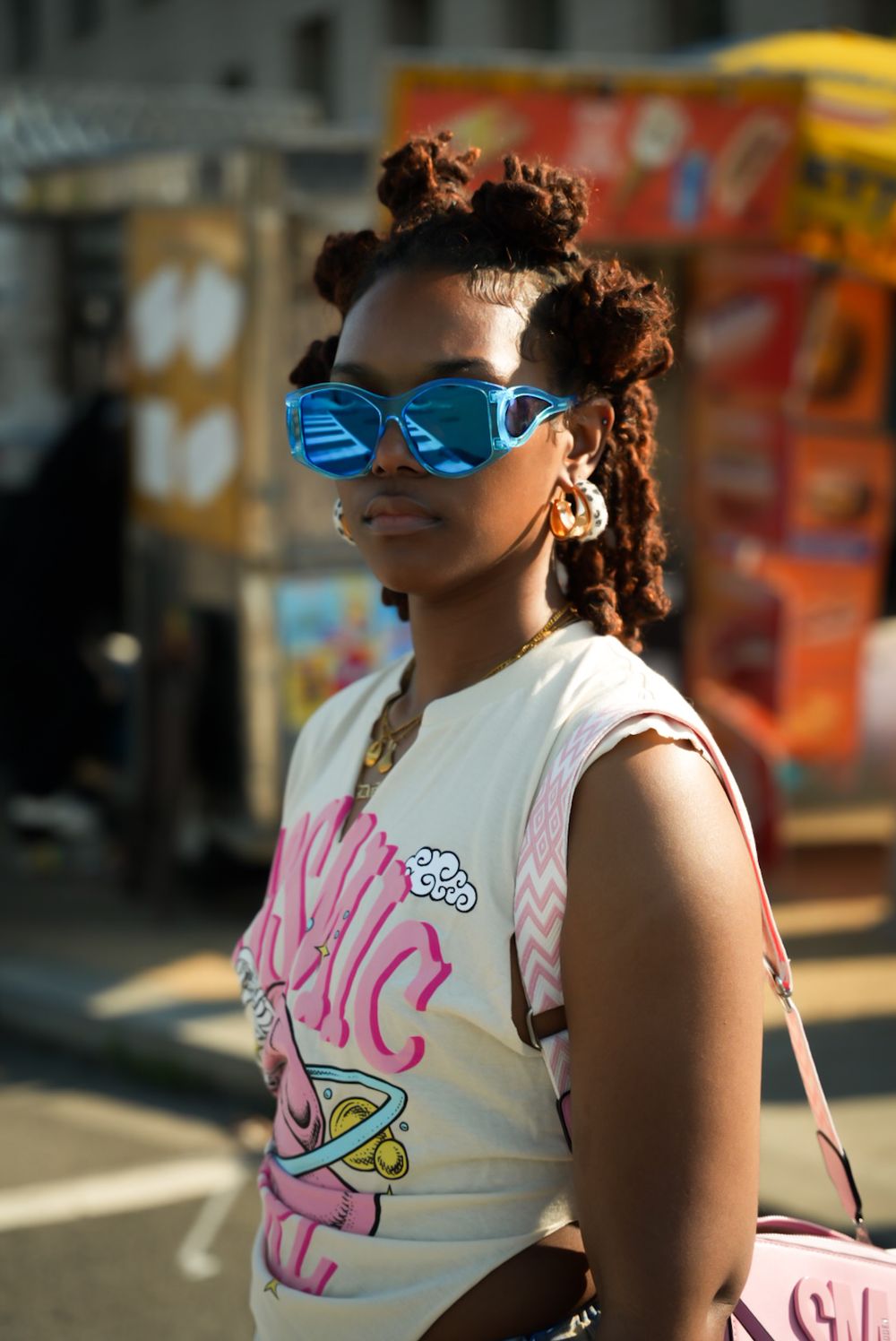 Person wearing blue sunglasses and a pink tank top stands in urban setting with blurred background.