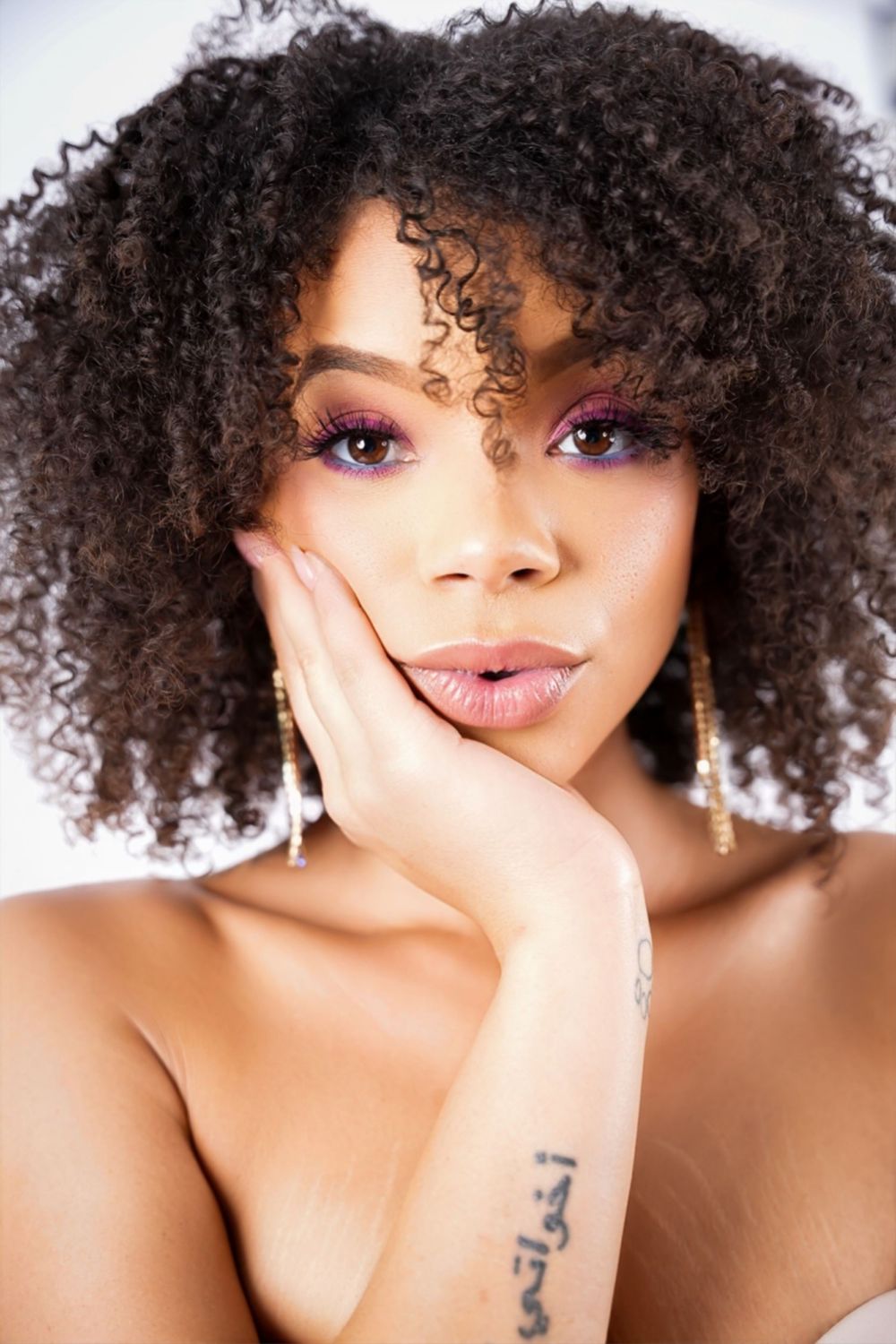 Beautiful portrait with natural curly hair, purple eye makeup, and soft lighting against white background.