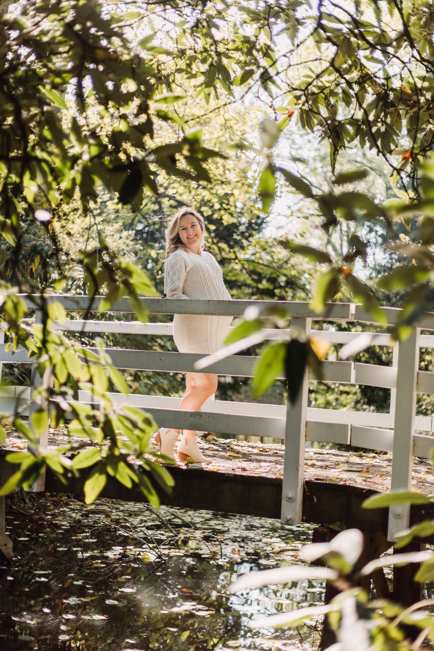 A pregnant woman sits peacefully on a wooden bench surrounded by dappled sunlight filtering through lush green foliage.