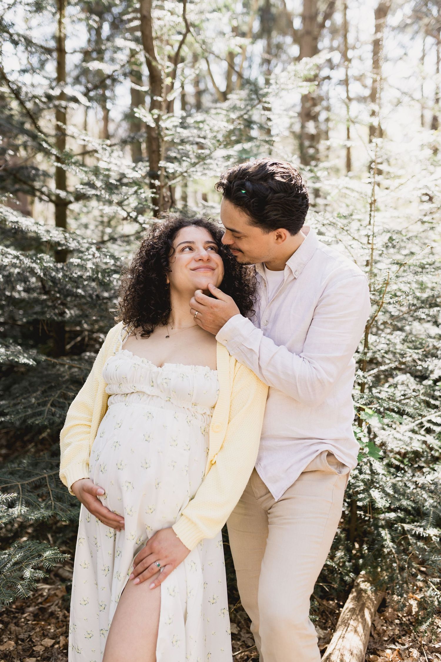 A couple shares intimate moments in a sun-dappled forest setting wearing coordinated white and cream outfits.
