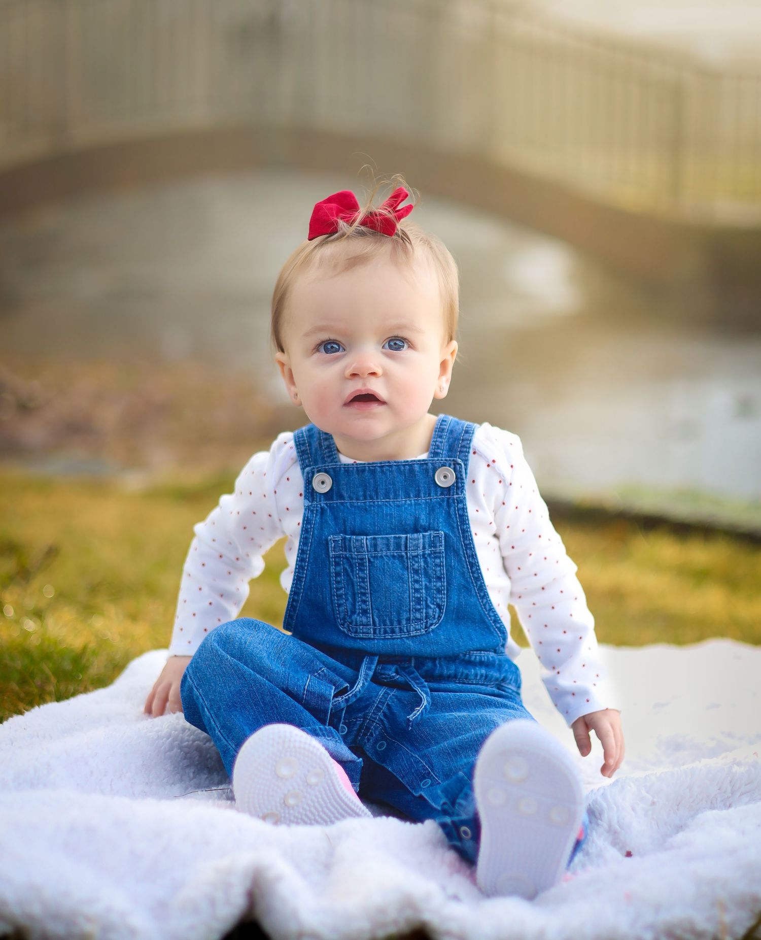 Baby in denim overalls and white top sitting on white blanket outdoors.