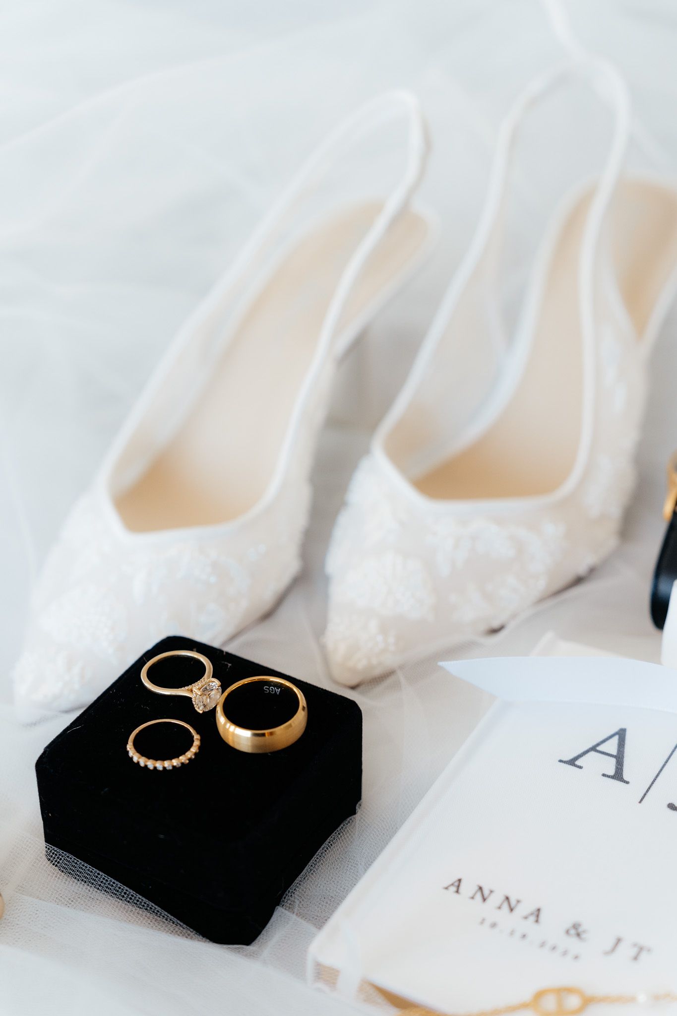White lace bridal shoes and gold wedding rings displayed on a black velvet ring box.