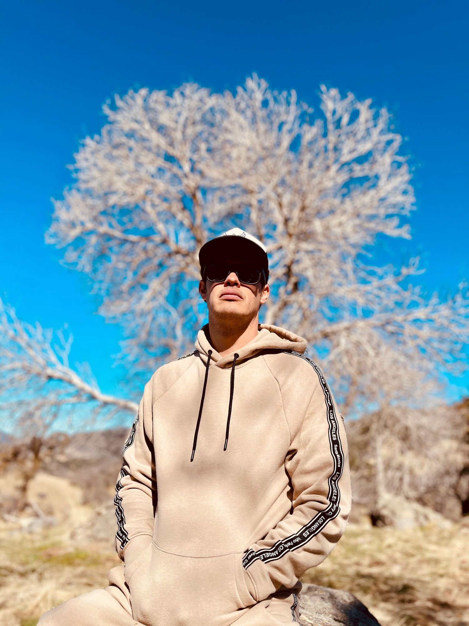 Figure in beige hoodie stands before frost-covered tree against bright blue sky.