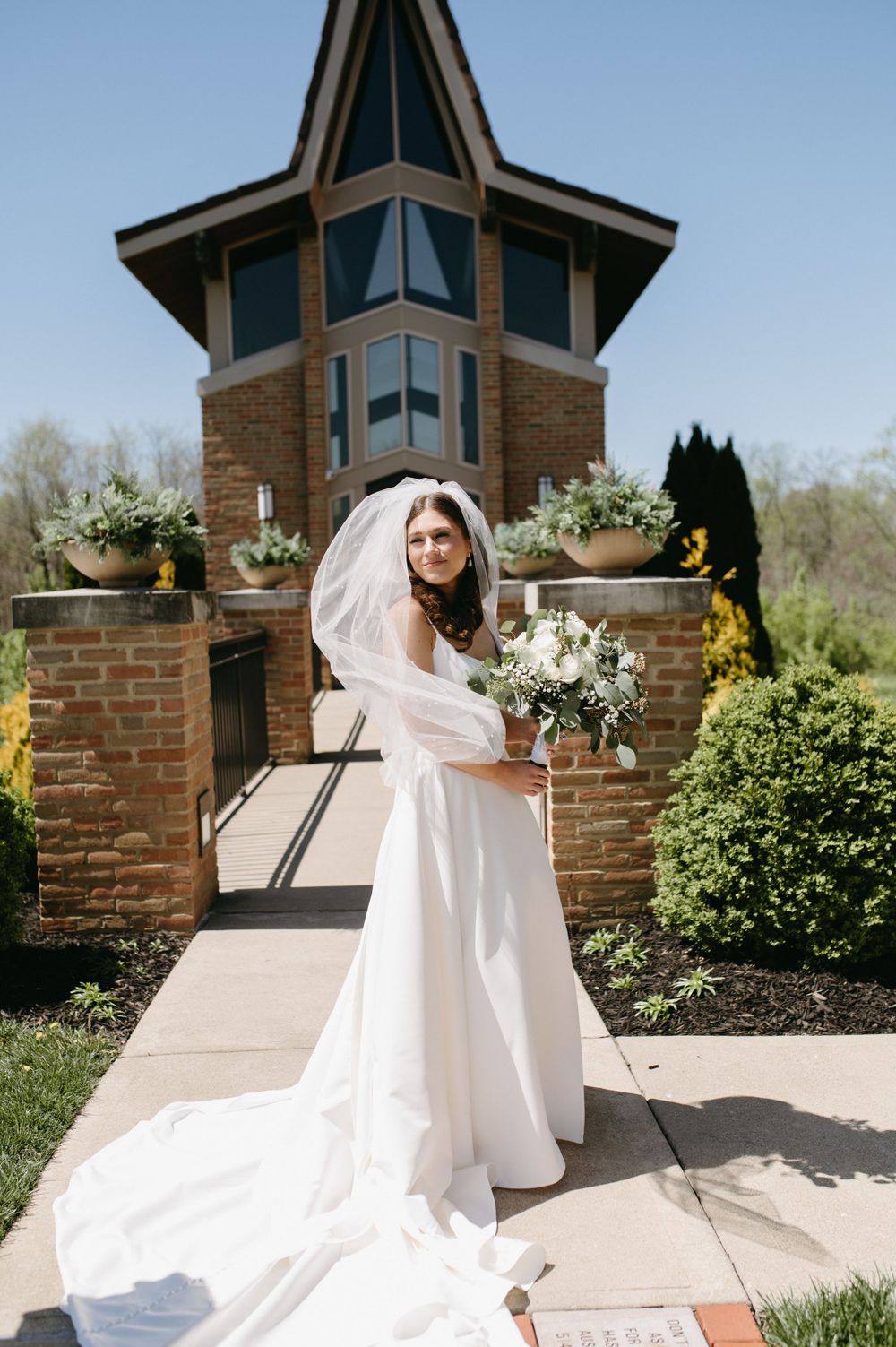 Bride in flowing white gown with veil holding bouquet in front of brick tower building.