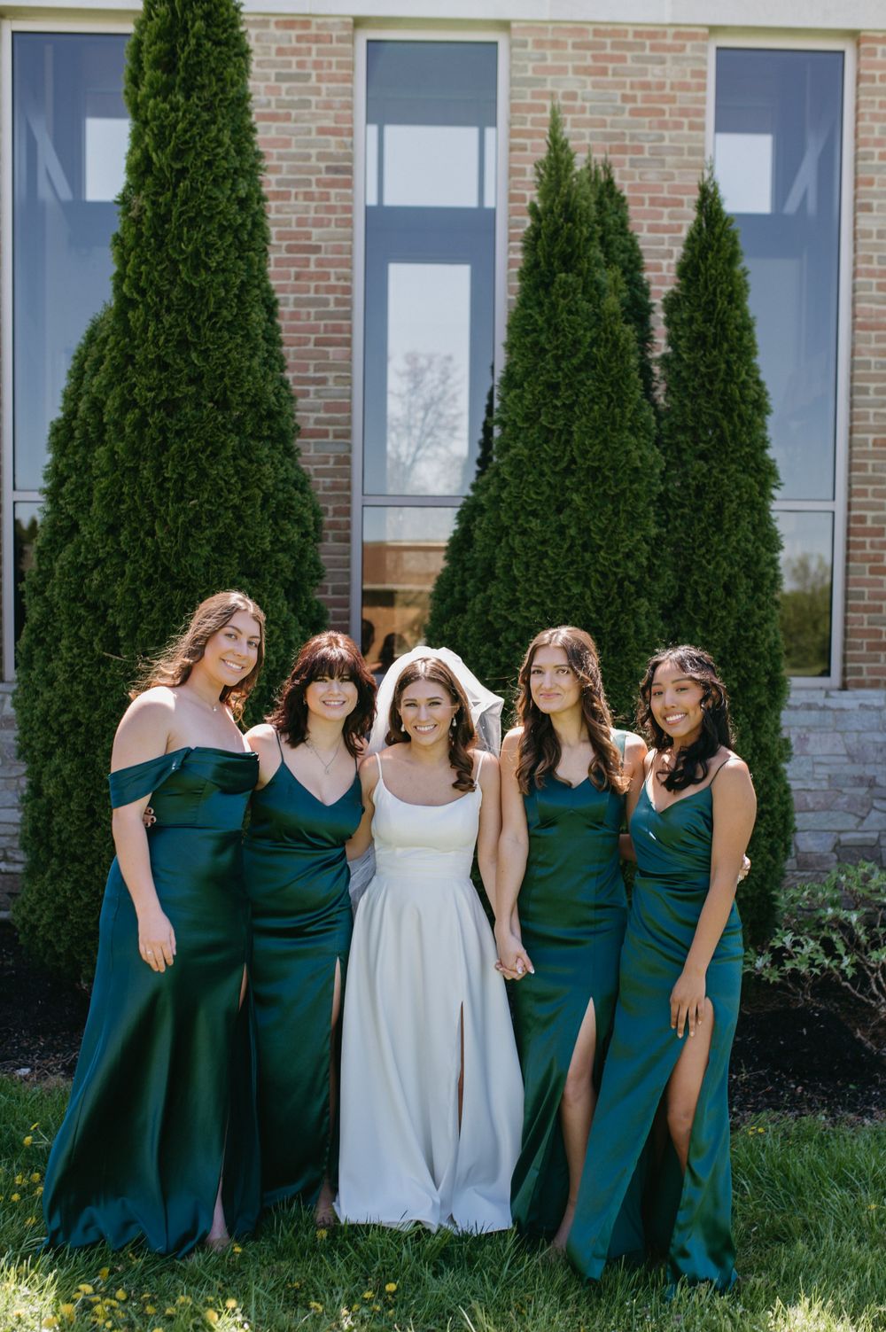 Bridal party posing together, bridesmaids wearing elegant emerald gowns against brick building and cypress trees.