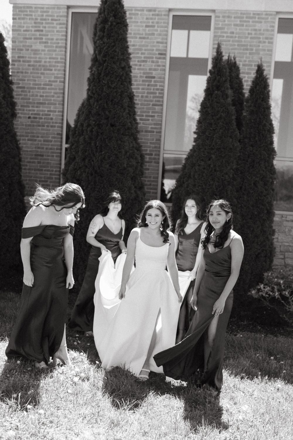 Black and white wedding photo showing bridesmaids helping with bride's dress near evergreen trees.