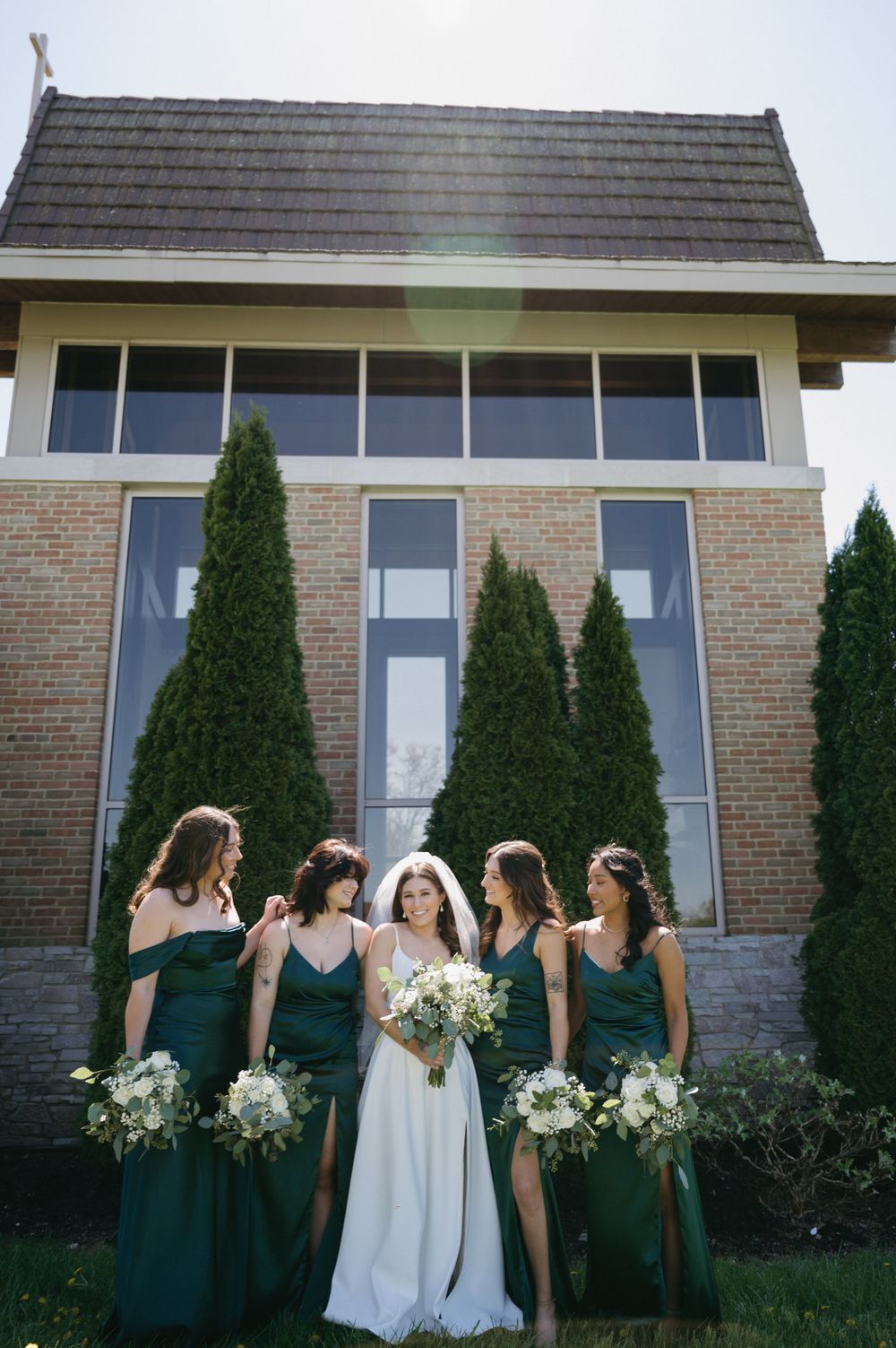 Wedding party with emerald green dresses and white bouquets posing in front of modern church building.