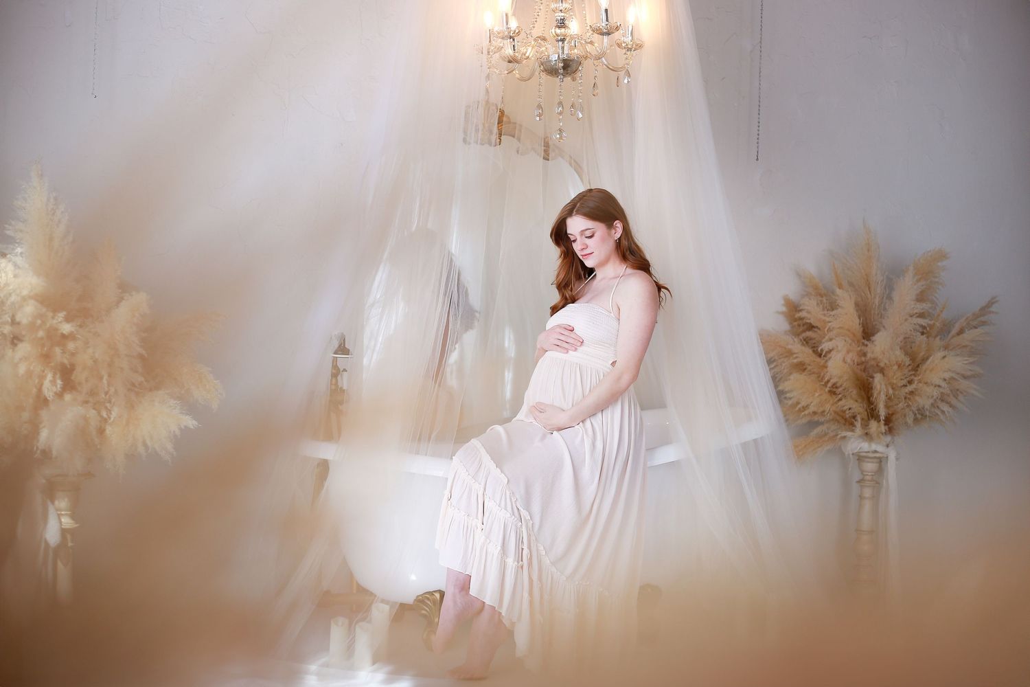A dreamy maternity photo with white flowy dress, crystal chandelier and pampas grass in a soft ethereal setting.