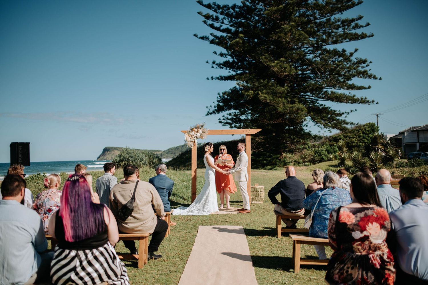 Wedding ceremony setup with wooden benches and arch against pine tree and ocean backdrop.