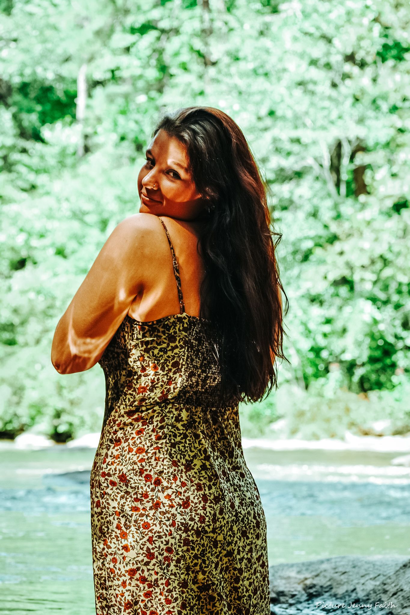 A person in a floral dress poses by a river in a lush green forest setting.