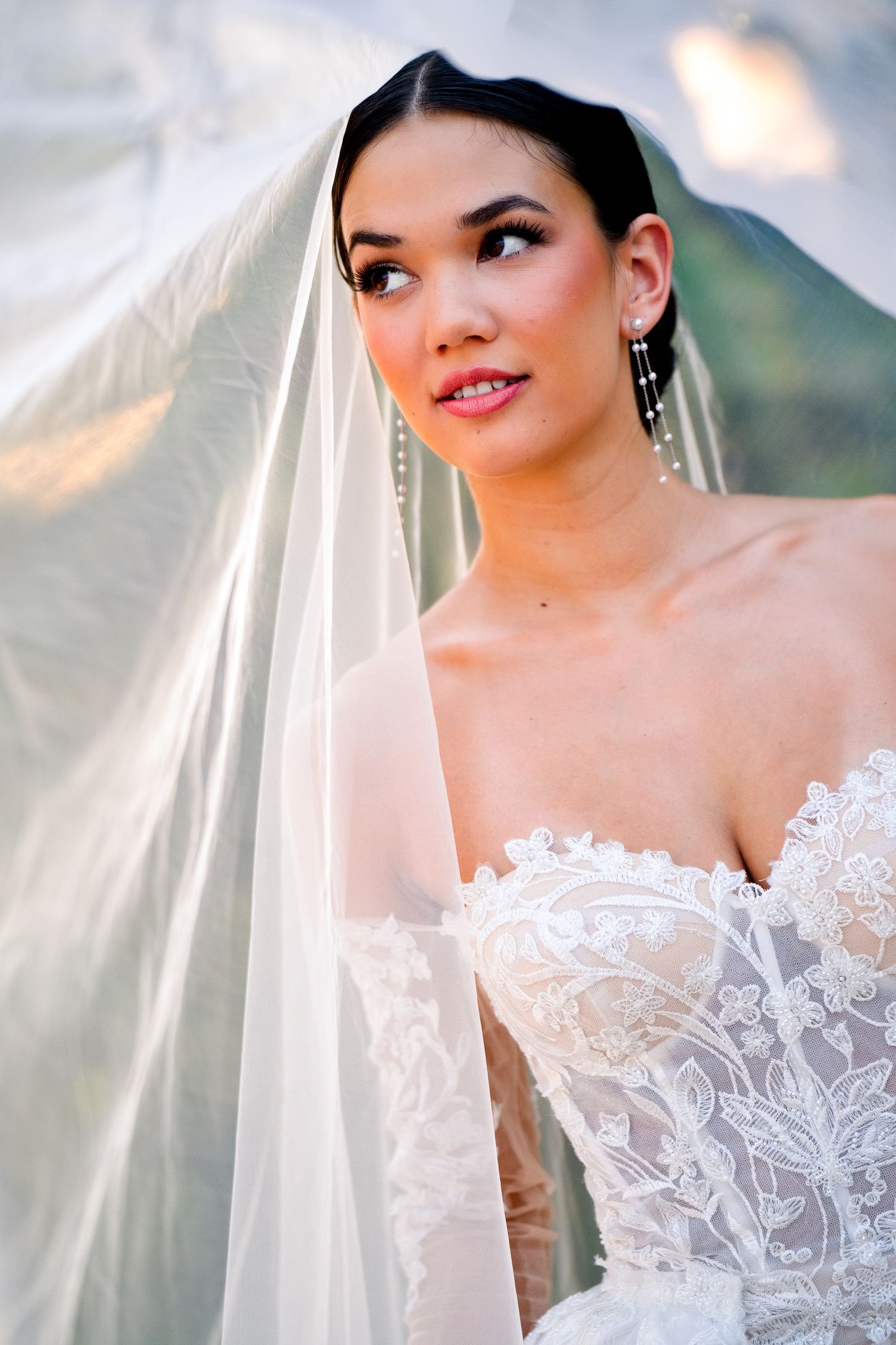 A bride in a white lace wedding dress with delicate details and a flowing veil poses in natural outdoor lighting.
