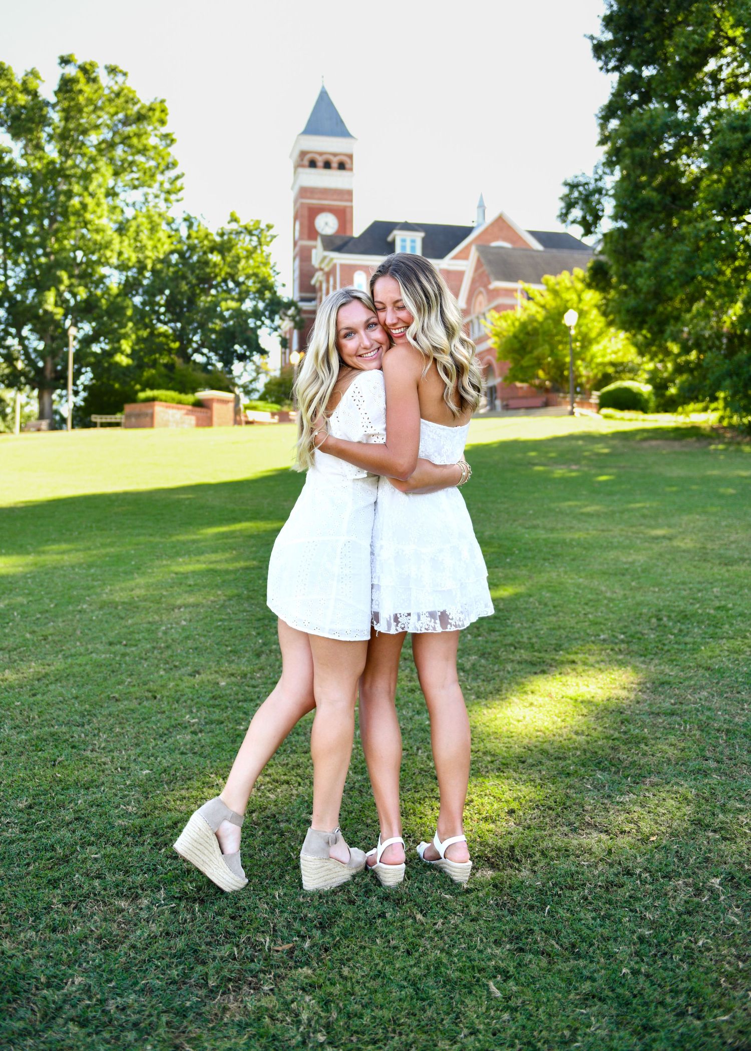 Two friends in white dresses embrace on sunny campus lawn with clock tower in background.
