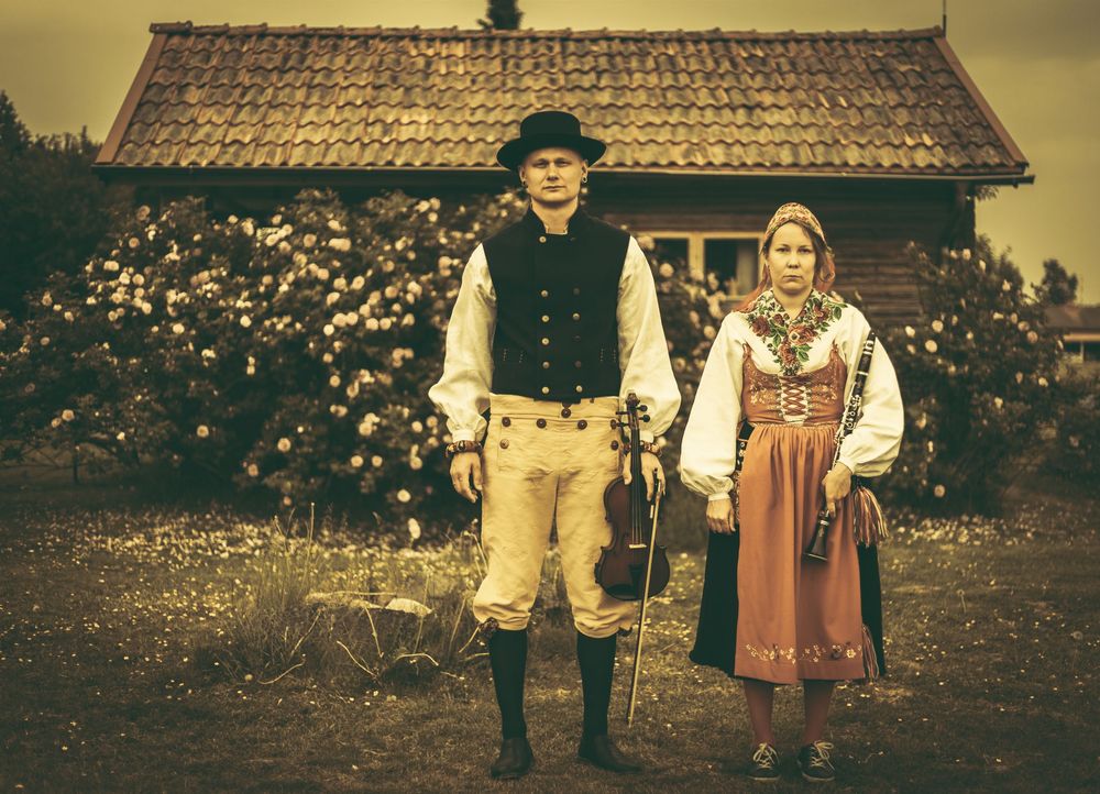 Par i traditionella folkdräkter med fiol i lantlig miljö med stuga. Gamaldags med sepia filter.