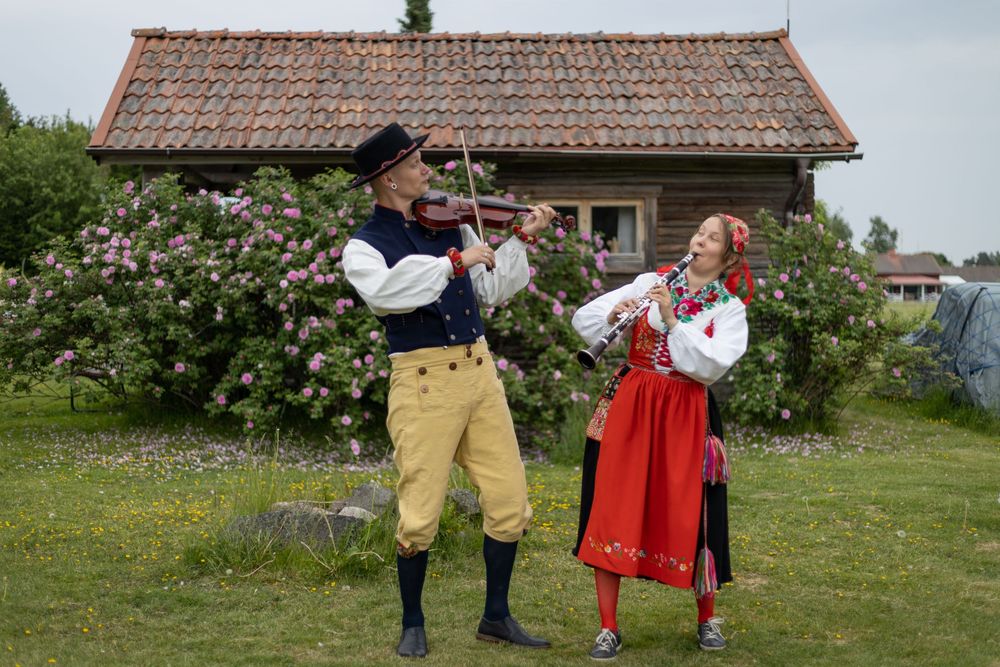 Par i traditionella folkdräkter med fiol i lantlig miljö med stuga i bakgrunden. Spelandes och glada.