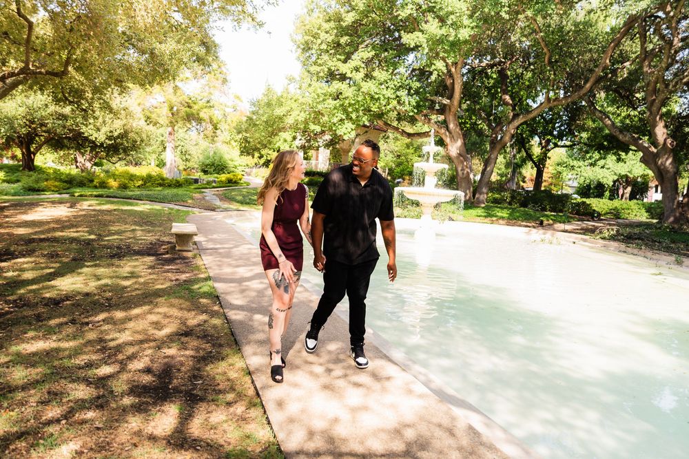 Couple walking hand in hand along a sunny park path beside a fountain, surrounded by lush green trees.
