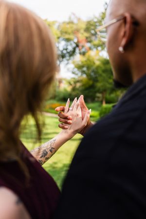 Couple holding hands outdoors showing engagement ring with tattoo visible, green garden background.