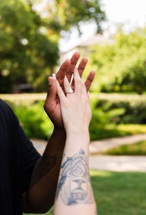 Two hands touching, one with a diamond engagement ring and the other with a tiger tattoo, outdoors in a garden.