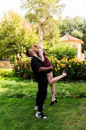 Couple sharing a romantic kiss in a lush park, man lifting woman near colorful flowers and a gazebo.