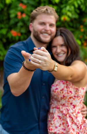 A couple in a blue shirt and floral dress show off engagement rings against a green garden backdrop with orange flowers. Engagement session