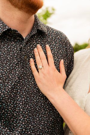 Close-up of a woman's hand with an elegant engagement ring resting on a man's dark floral button-up shirt. irving engagement session